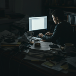 A disorganized wooden desk overflowing with stacks of binders, papers, and tangled electronic cables.