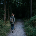 Woman in green jacket walking on dirt path through dense forest