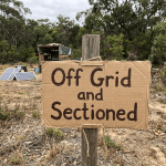 Cardboard sign reading 'Off Grid and Sectioned' tied to a wooden post near solar panels and a small shelter in a wooded area